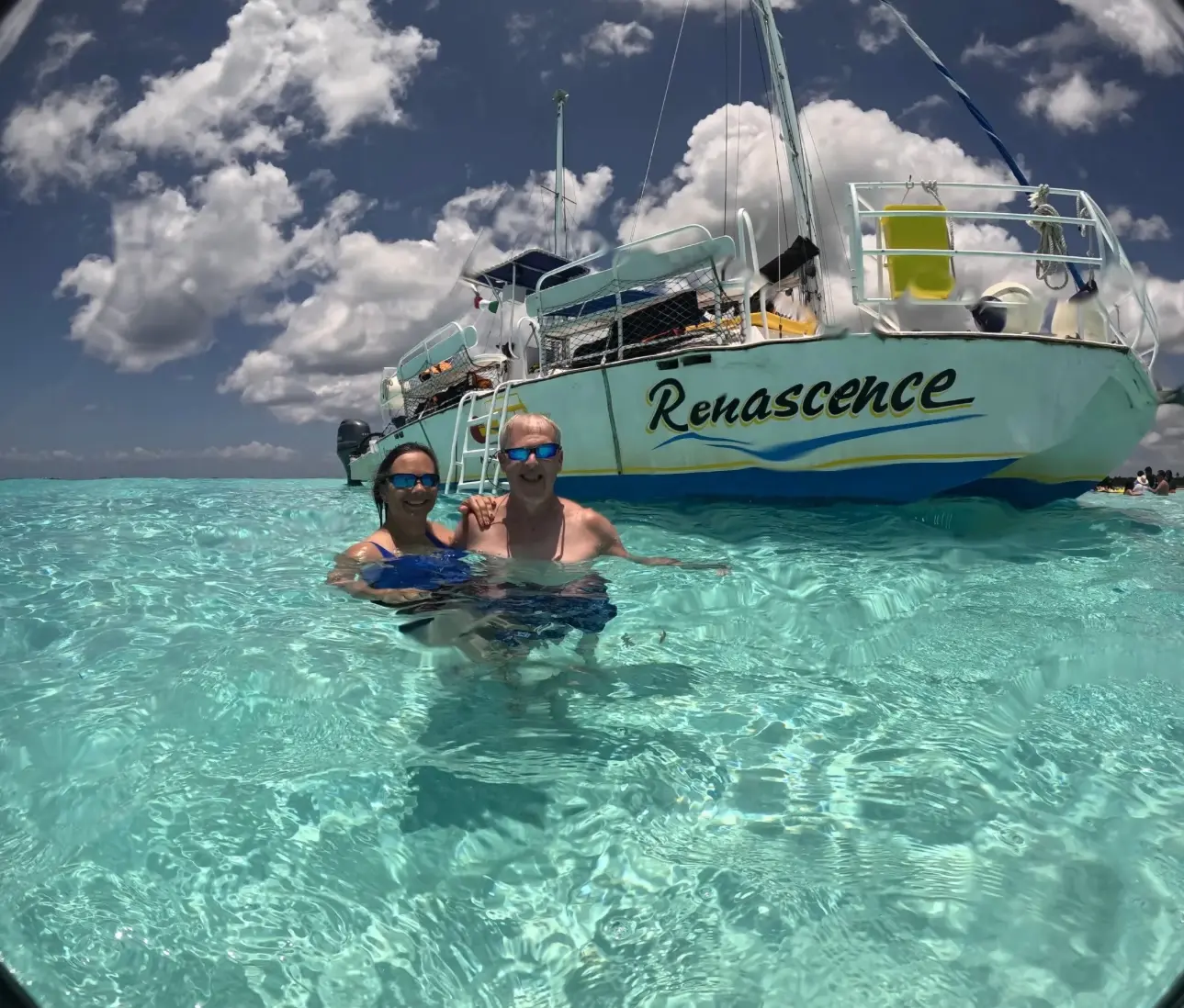 A couple stands waist-deep in clear turquoise water in front of the sailing catamaran at El Cielo