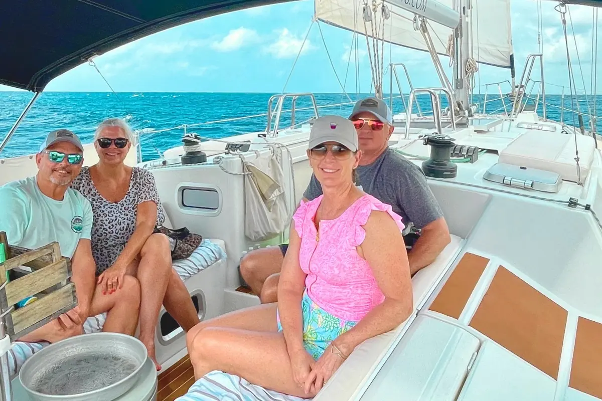 Four guests in caps and sunglasses seated in the cockpit of a sailing yacht on a sunny sail