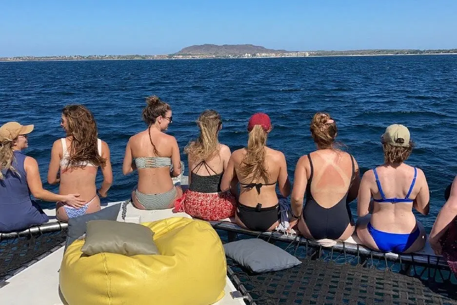 Guests sit in a row along the catamaran's bow, looking across the bay toward the distant shore