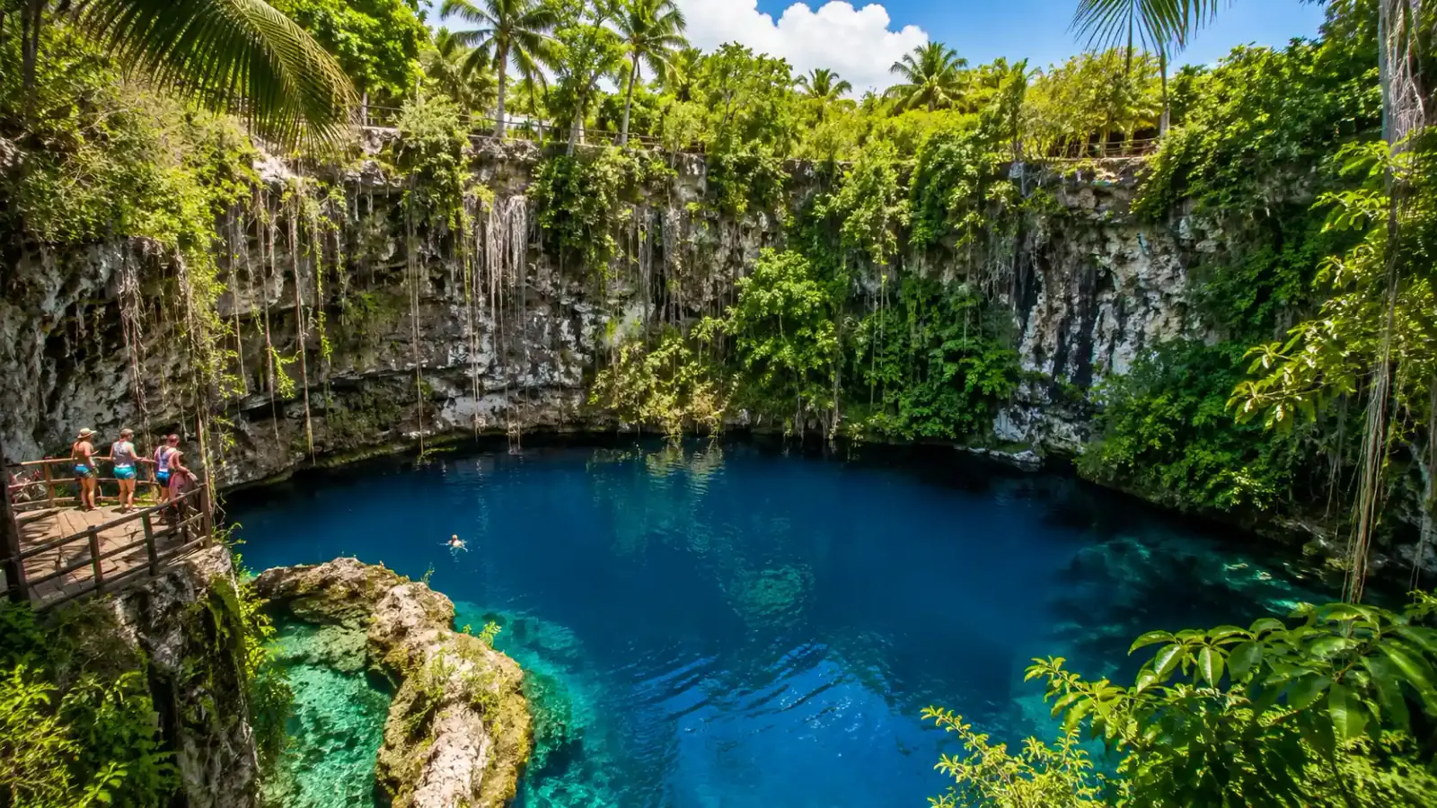 Clear blue Hoyo Azul cenote surrounded by limestone cliffs, tropical greenery, wooden viewing deck, and visitors enjoying an inland Cap Cana excursion.