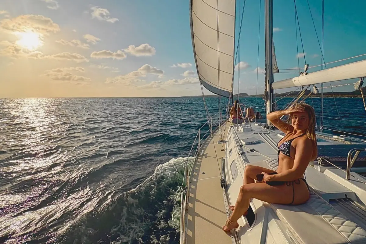 Woman in a bikini sitting on the side deck as the sailing yacht heels under sail off Curaçao at golden hour