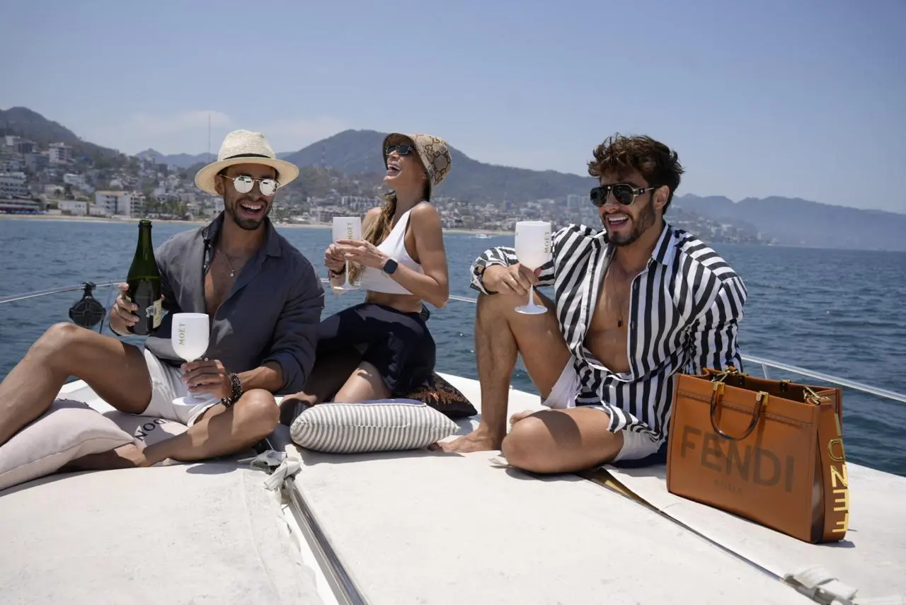 Three friends laugh and raise their glasses on the yacht's sunny bow above the bay