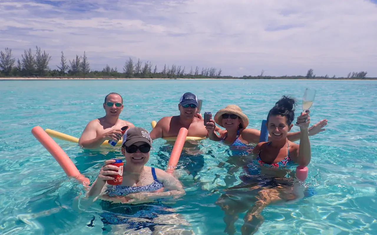 Friends with pool noodles raising drinks in clear shallow water off a tree-lined Riviera Maya shore