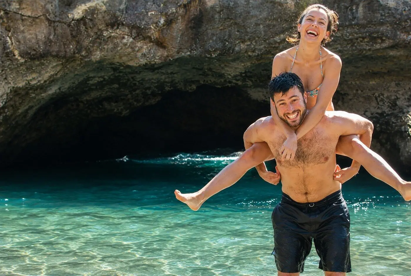 Man giving a laughing woman a piggyback ride in clear turquoise water beside a sea cave near Puerto Vallarta