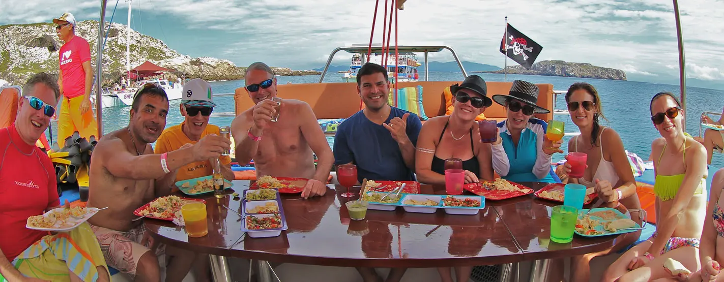 Guests share plates of food and colorful drinks around a table on the catamaran's deck