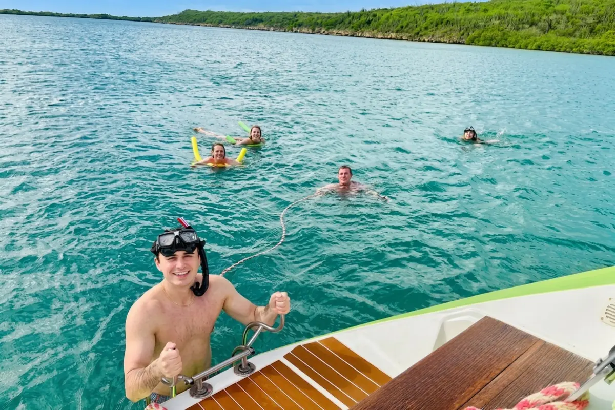 Man in a snorkel mask climbing a yacht's swim ladder as friends float on noodles in a Curaçao bay