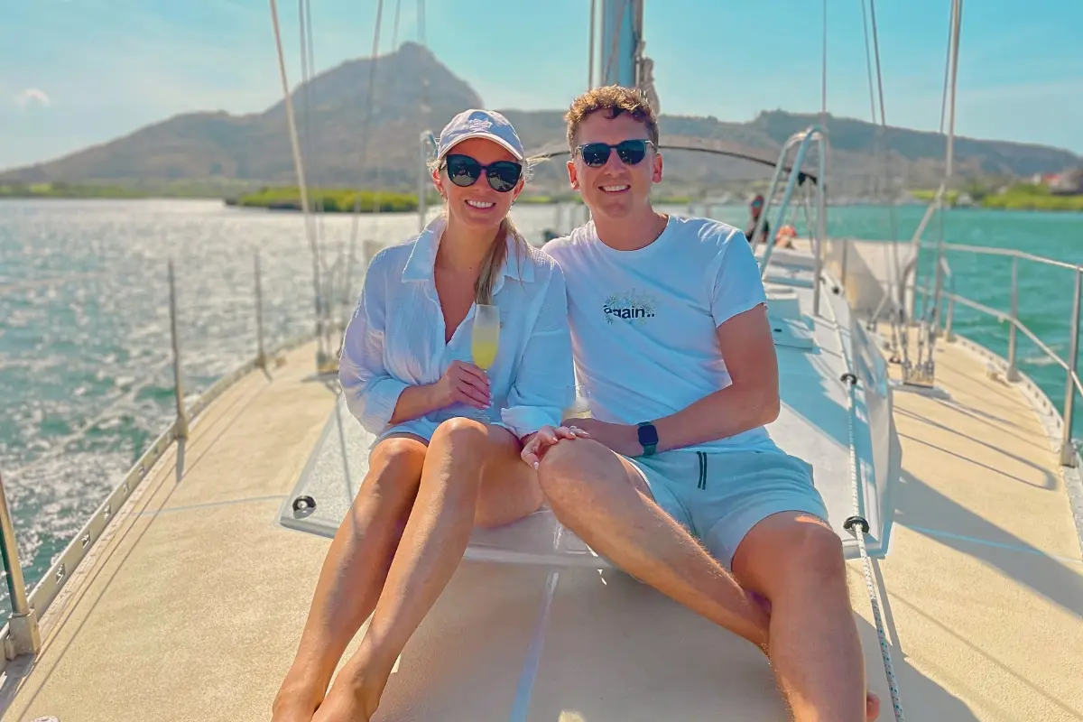 Couple in sunglasses sitting on the sailing yacht's deck with drinks, a hill rising behind them in Curaçao