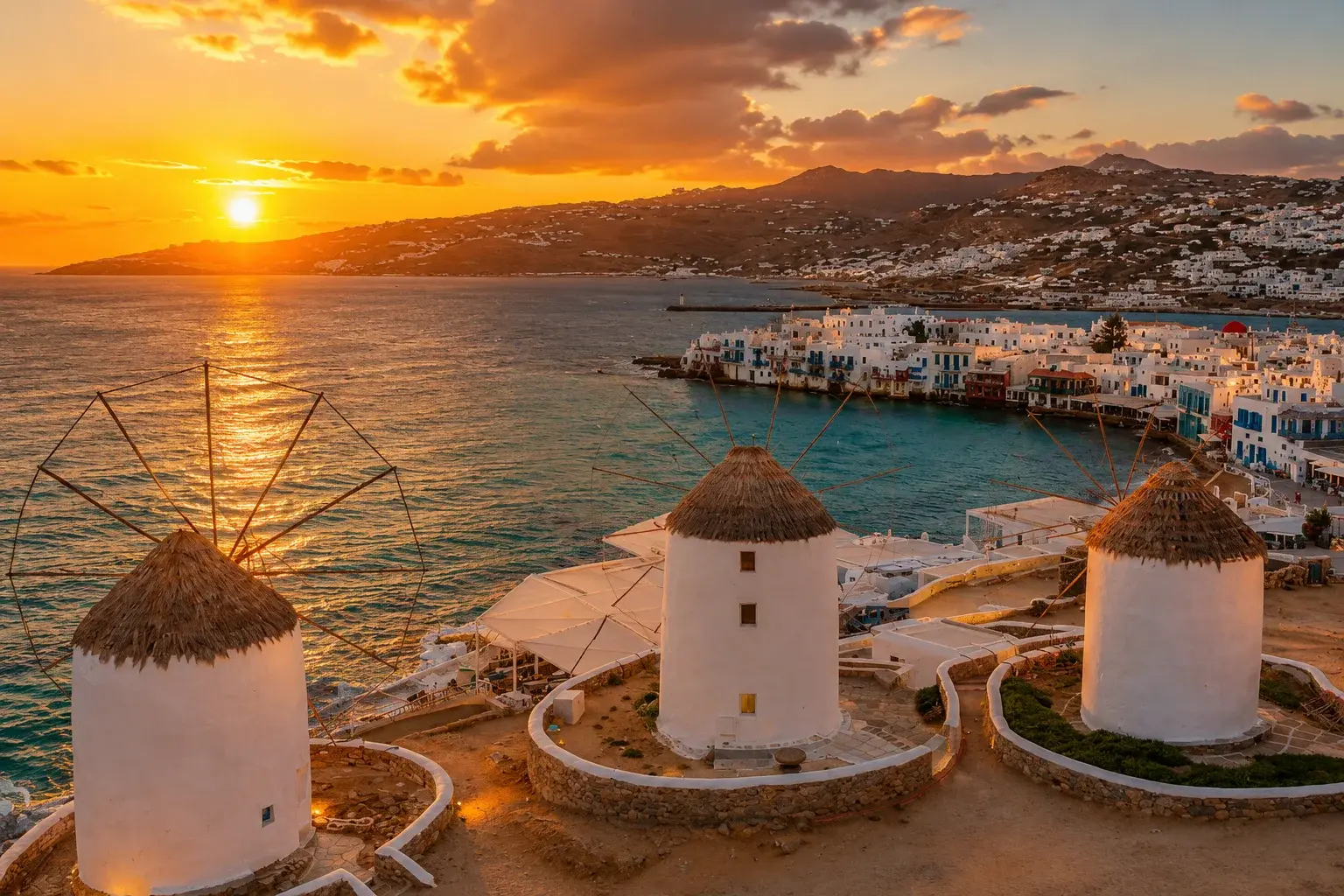 Sunset view of Mykonos with iconic white windmills overlooking the blue Aegean Sea, whitewashed coastal buildings, and surrounding hills.