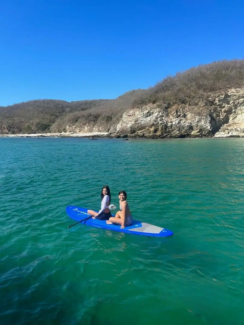 Two women sitting on a blue paddleboard in calm green water below a rocky headland on Banderas Bay