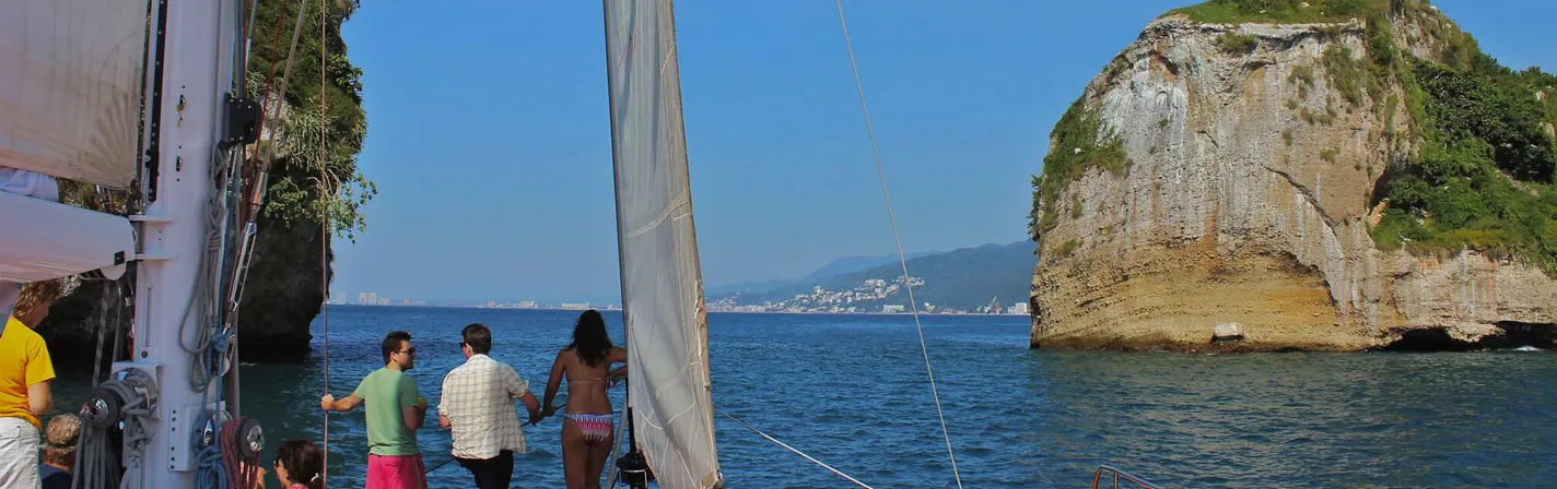 Guests at the bow of the sailing catamaran looking toward a rocky islet and the distant Puerto Vallarta shore