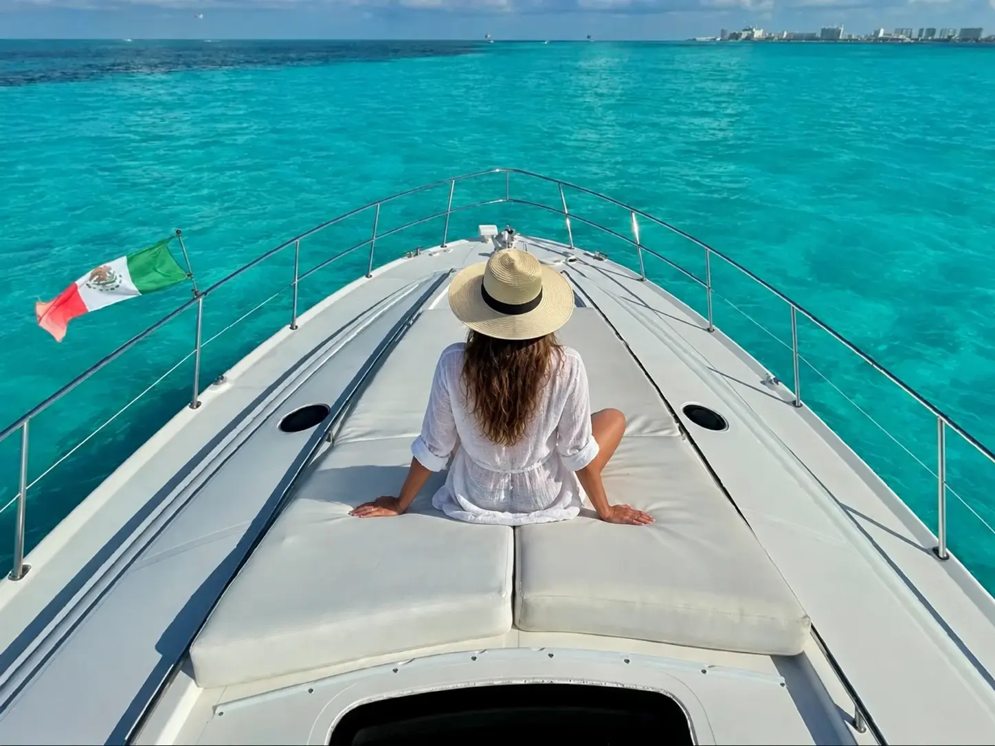 Woman in a sun hat sitting on the bow of a yacht, looking ahead over turquoise water to the Cancún shoreline