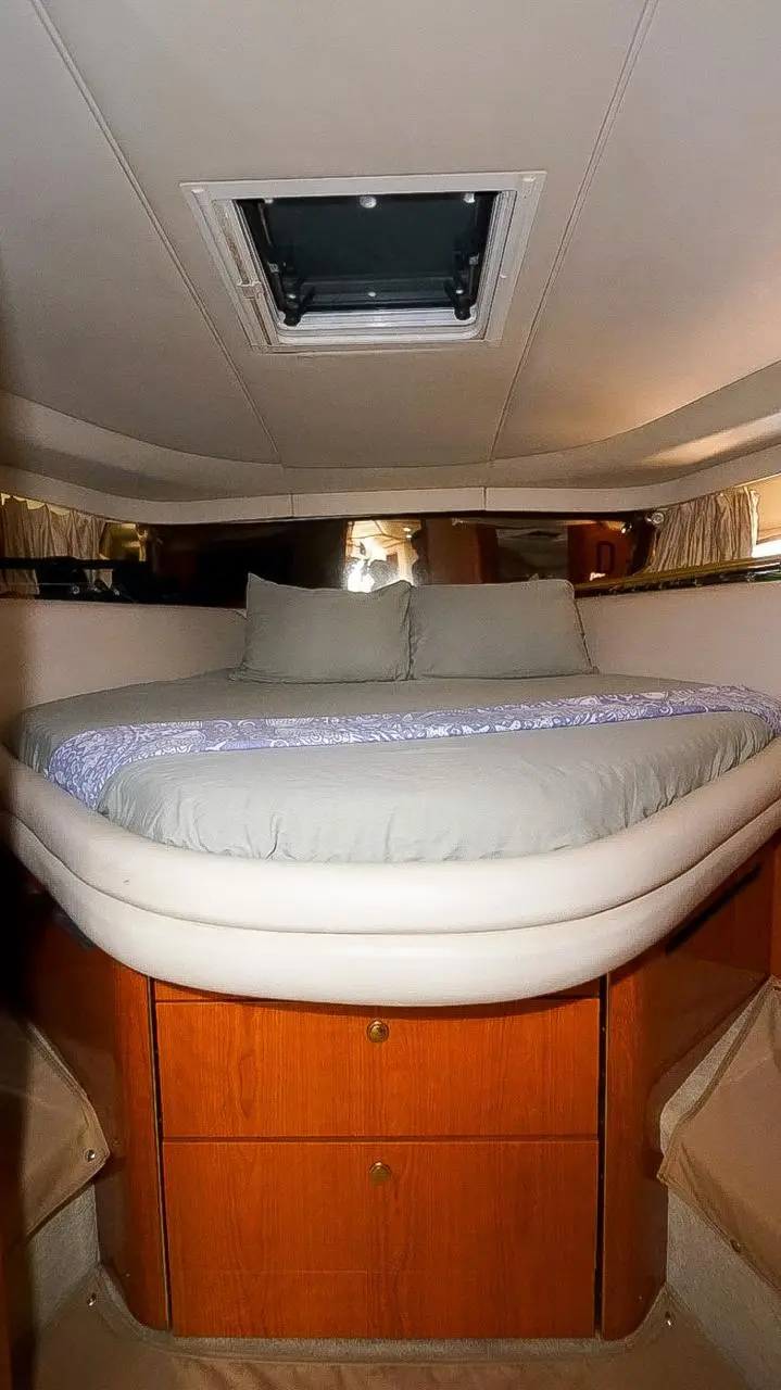 Cabin with a double bed in white linens beneath an overhead hatch, wood drawers below