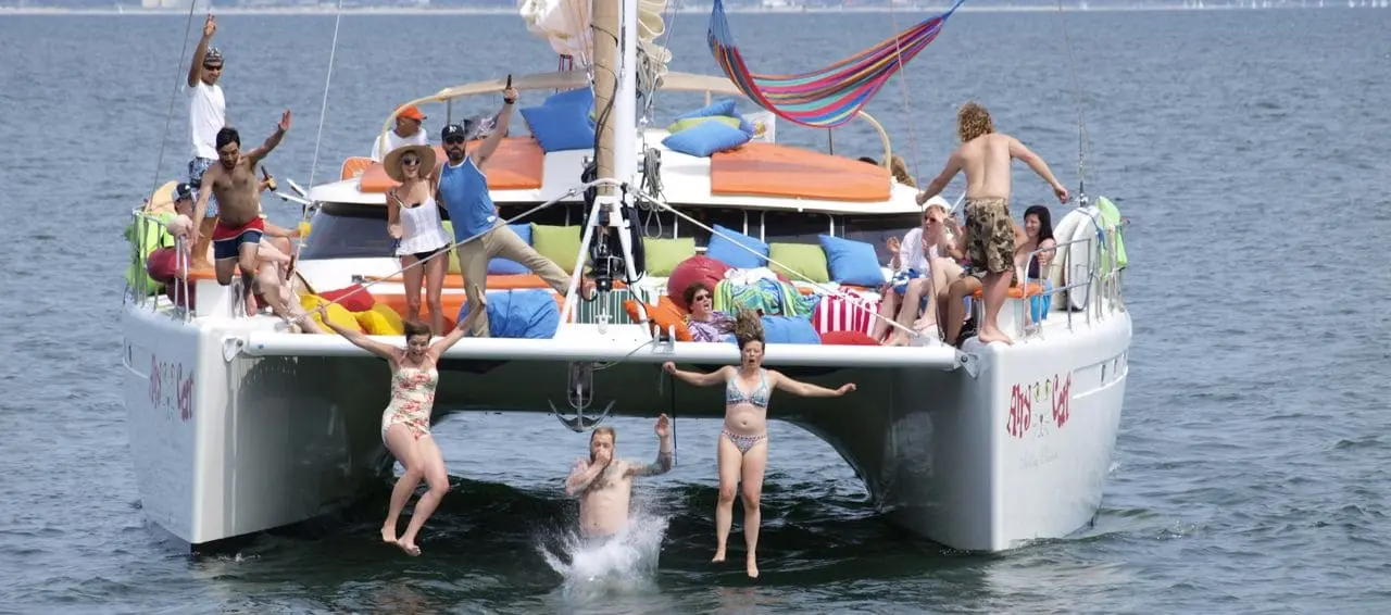 Guests jump from the front of a sailing catamaran while others wave from its cushioned deck
