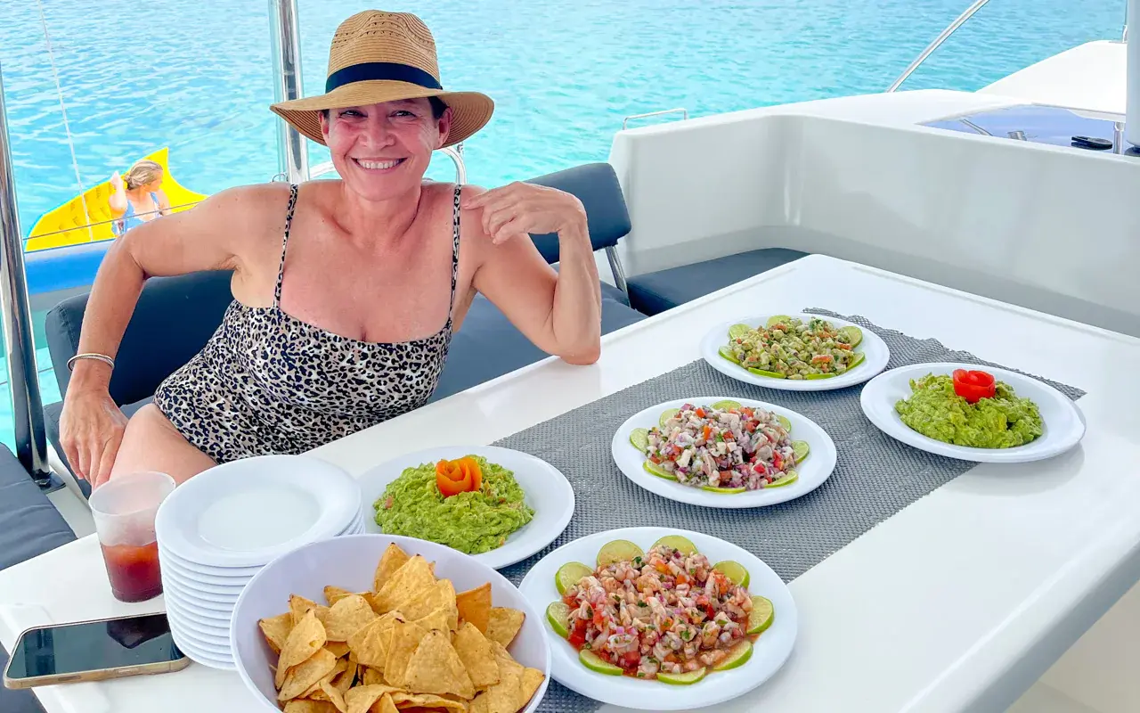 Smiling woman in a sun hat at the catamaran's cockpit table, set with ceviche, guacamole and chips