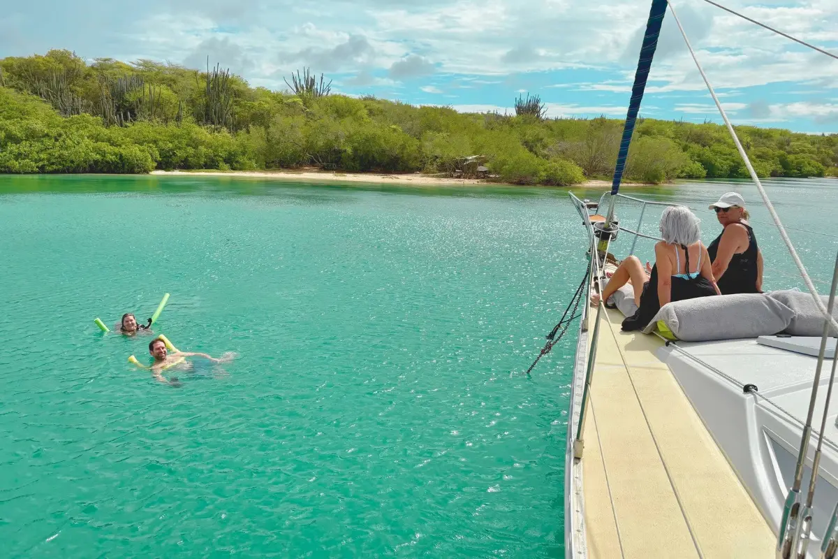 Two women sitting on a sailing yacht's side deck watching swimmers in a turquoise Curaçao bay