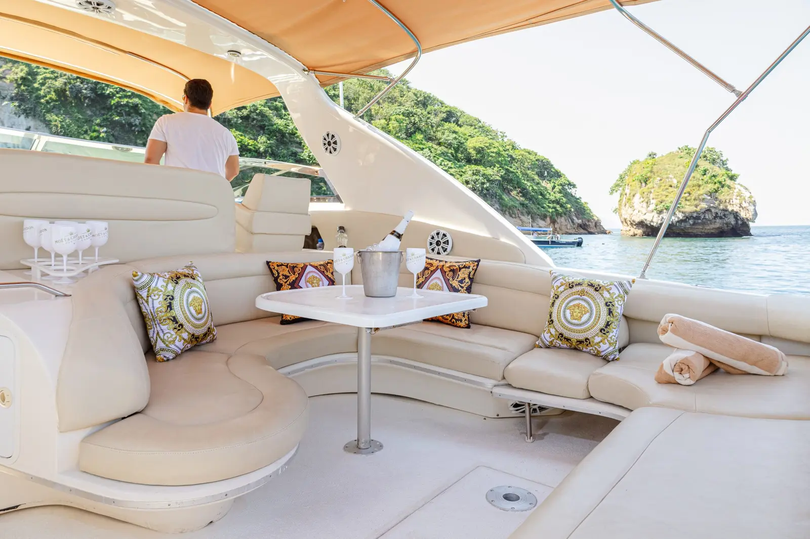 The yacht's shaded cockpit lounge with cream seating, glasses on the table and a man at the helm
