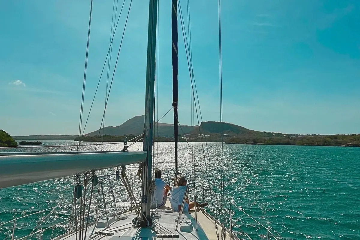 Two guests relaxing on the bow of a sailing yacht gliding across calm water toward Curaçao's green hills