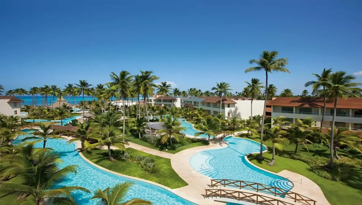 Winding lagoon pools with wooden footbridges among palm trees at a Punta Cana resort, the sea in the distance