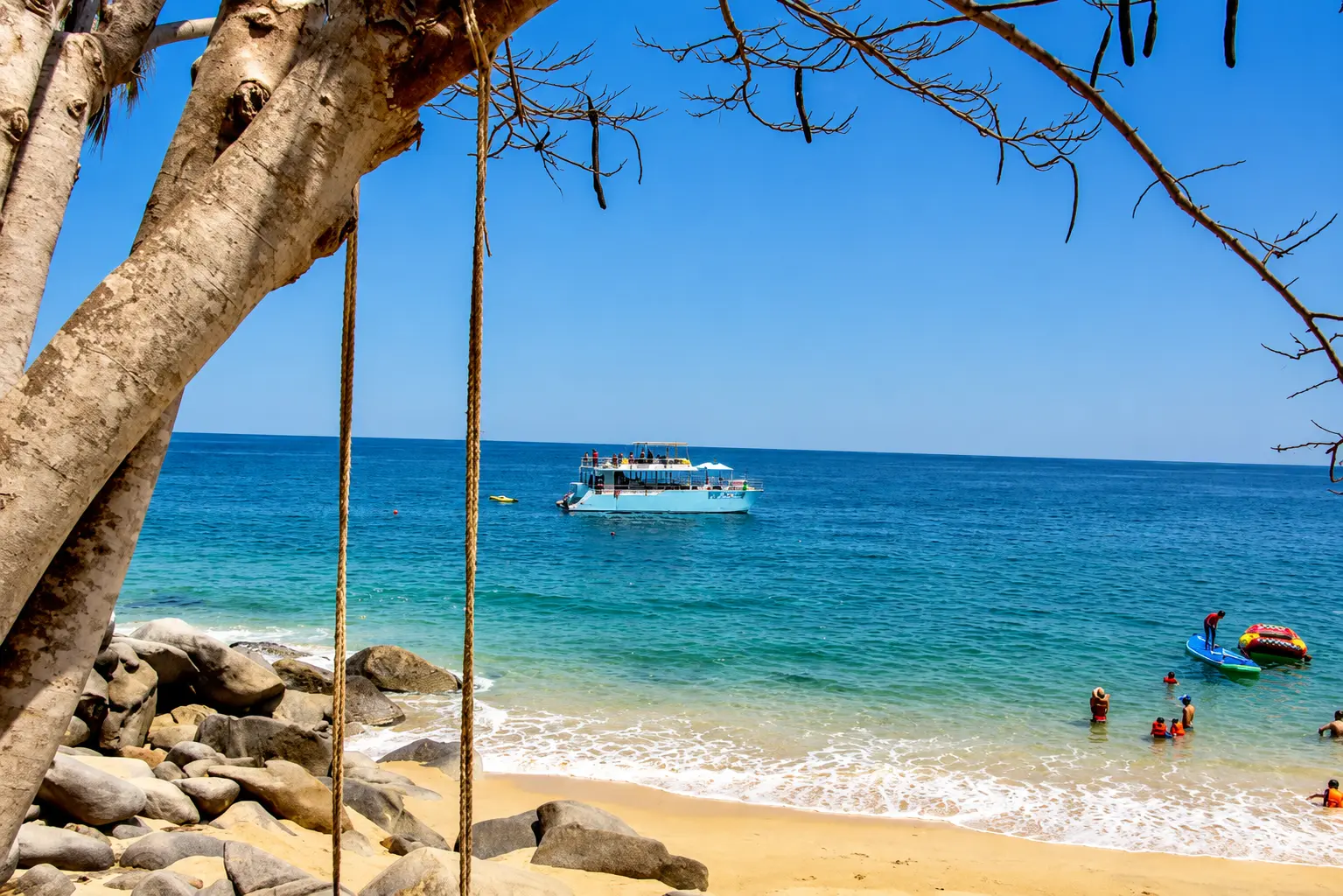 Catamaran anchored off a tree-shaded beach near Puerto Vallarta as guests swim and kayak by the shore