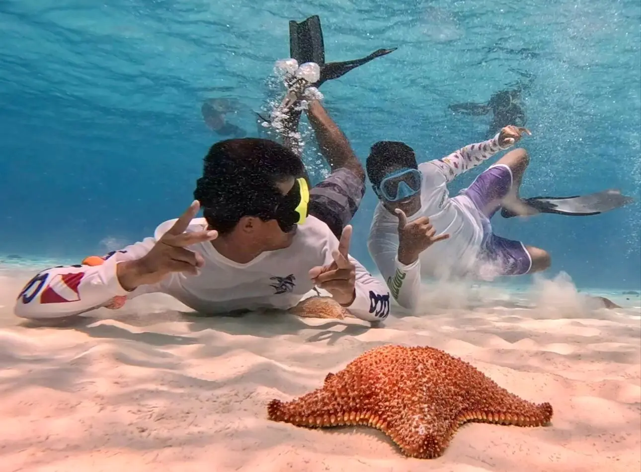 Two snorkelers in rash guards posing with hand signs on the sandy seabed behind an orange starfish