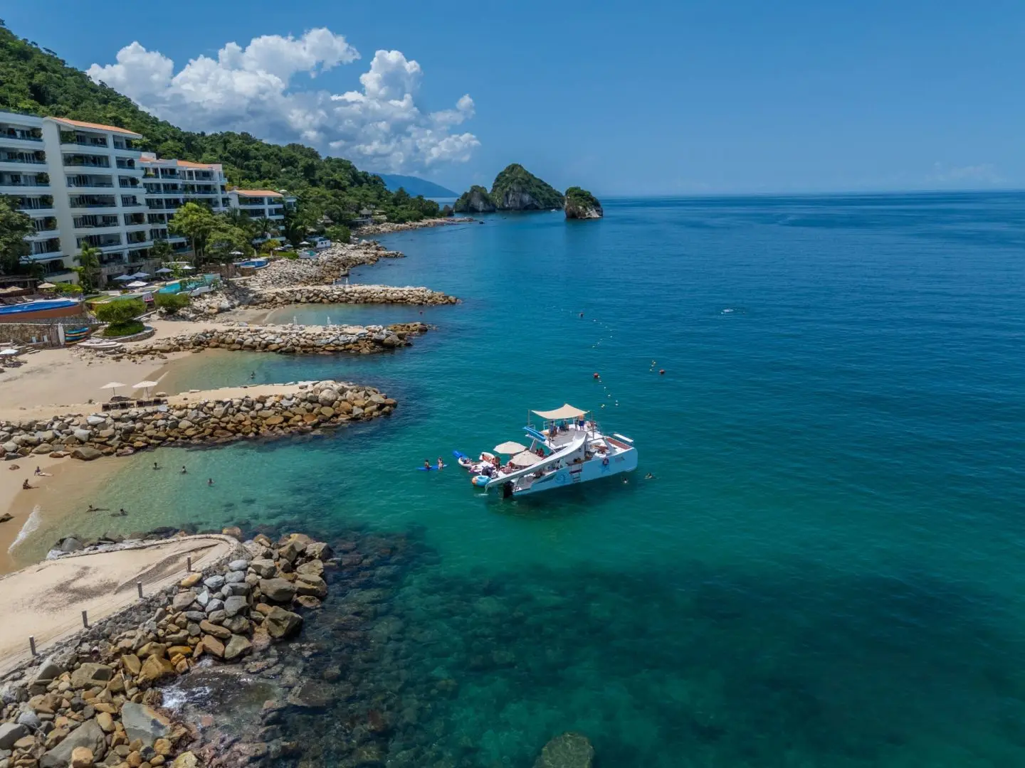 Aerial view of a catamaran anchored off a hotel-lined beach with rock jetties in Puerto Vallarta