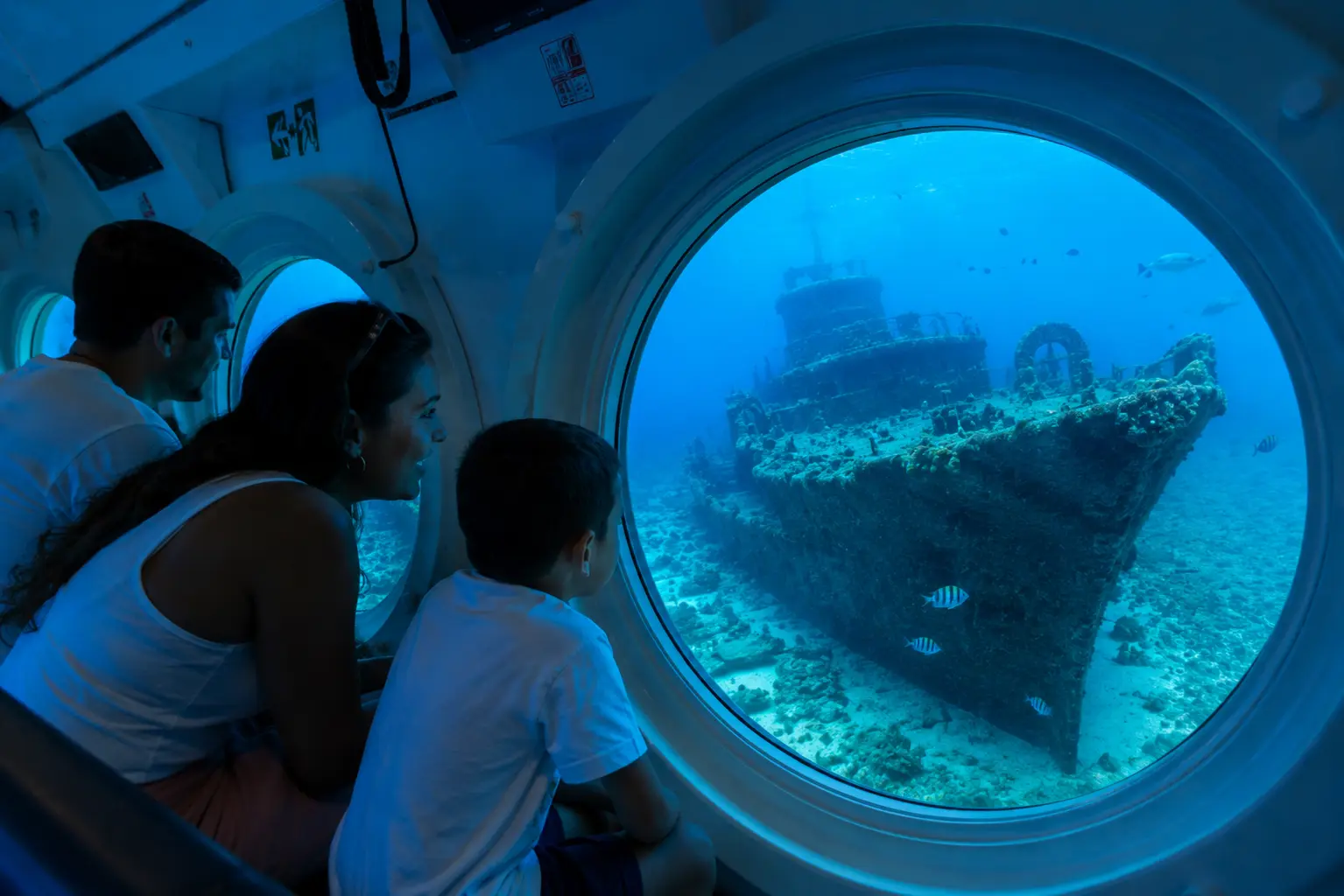 A family peers through a submarine porthole at a sunken shipwreck on the sea floor