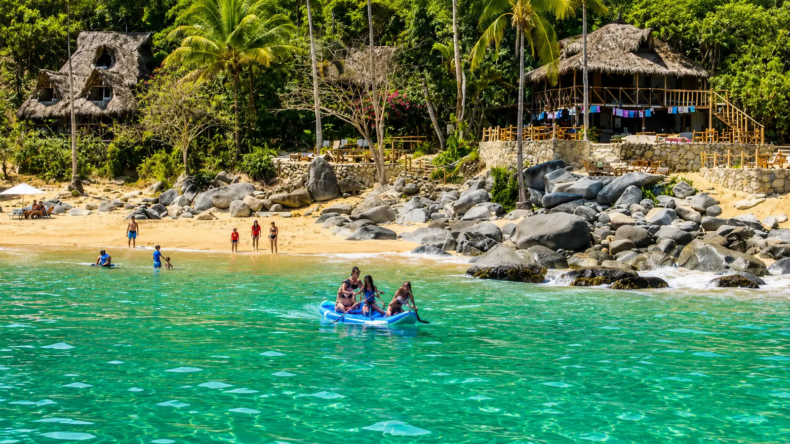 Guests ride a small motorboat toward a palm-fringed beach with thatched huts near Puerto Vallarta