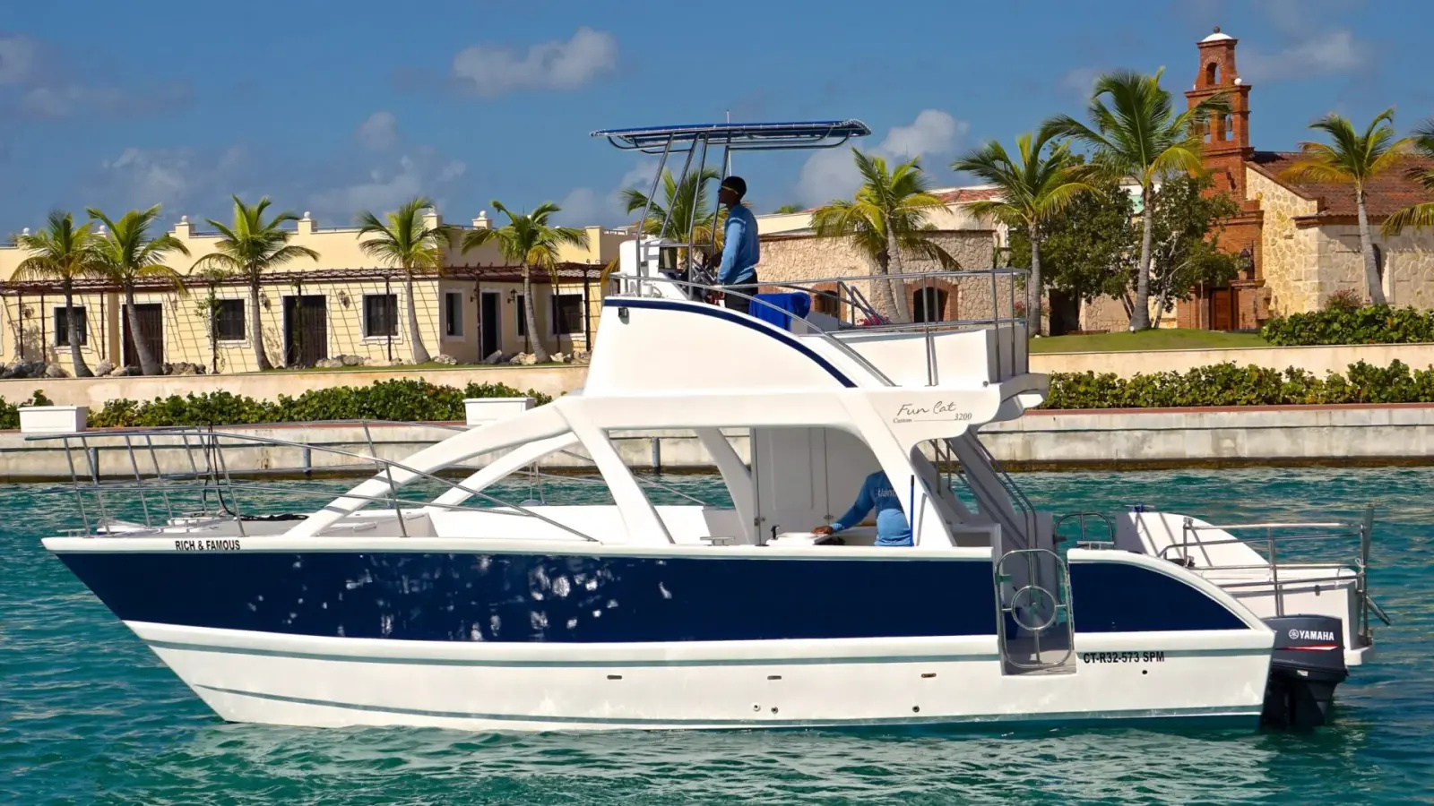 White catamaran cruise boat cruising on turquoise water near a palm-lined resort with a Mediterranean-style waterfront building in the background.