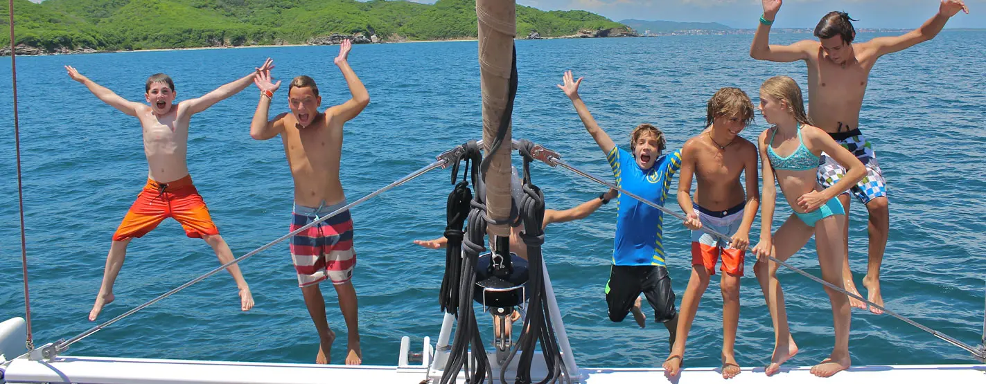 Kids leap into the air on the catamaran's bow with the bay and green hills behind