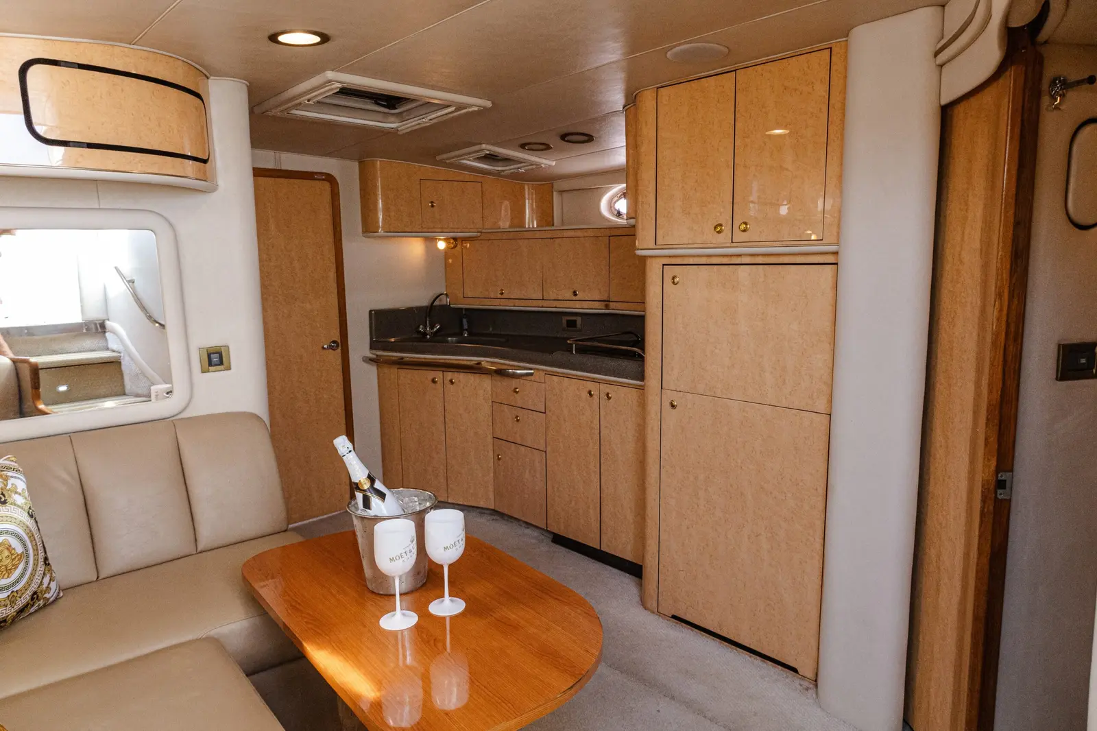Yacht salon with a cream sofa, champagne on ice on the wooden table and a wood-paneled galley
