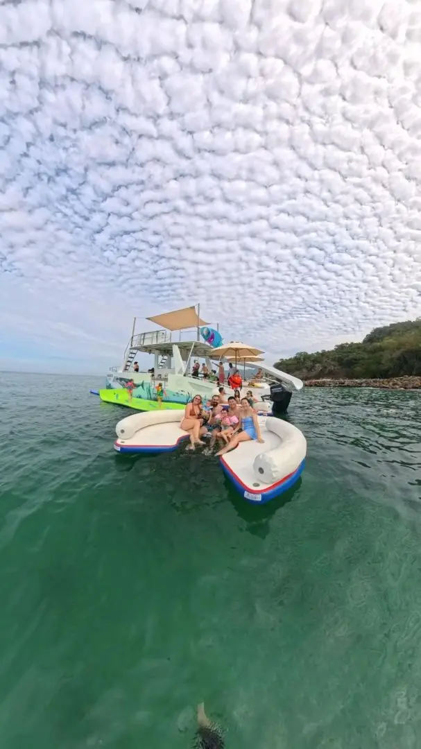 Wide-angle view of guests on an inflatable platform beside the catamaran under a rippled cloud sky