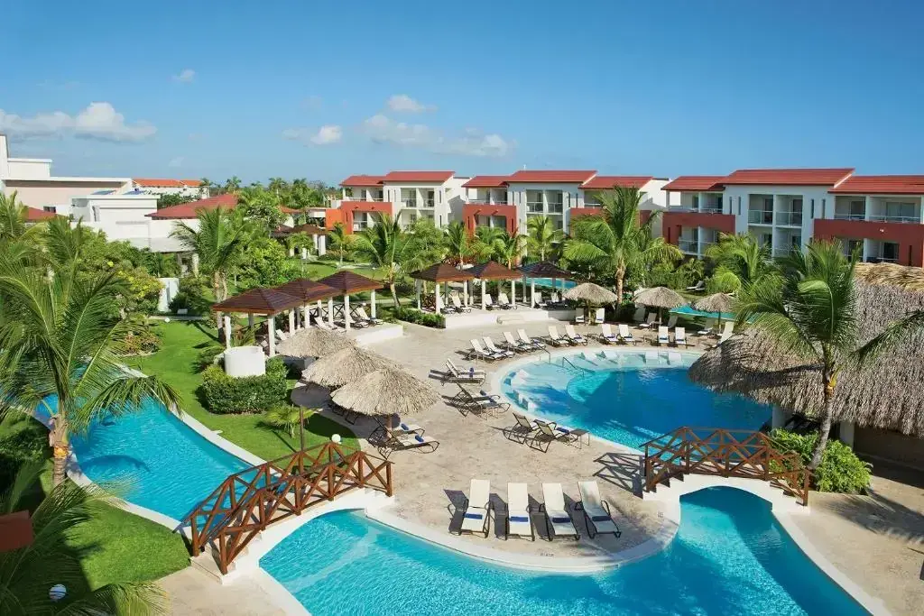Resort pools with wooden footbridges, thatched umbrellas and loungers beside red-roofed buildings in Punta Cana
