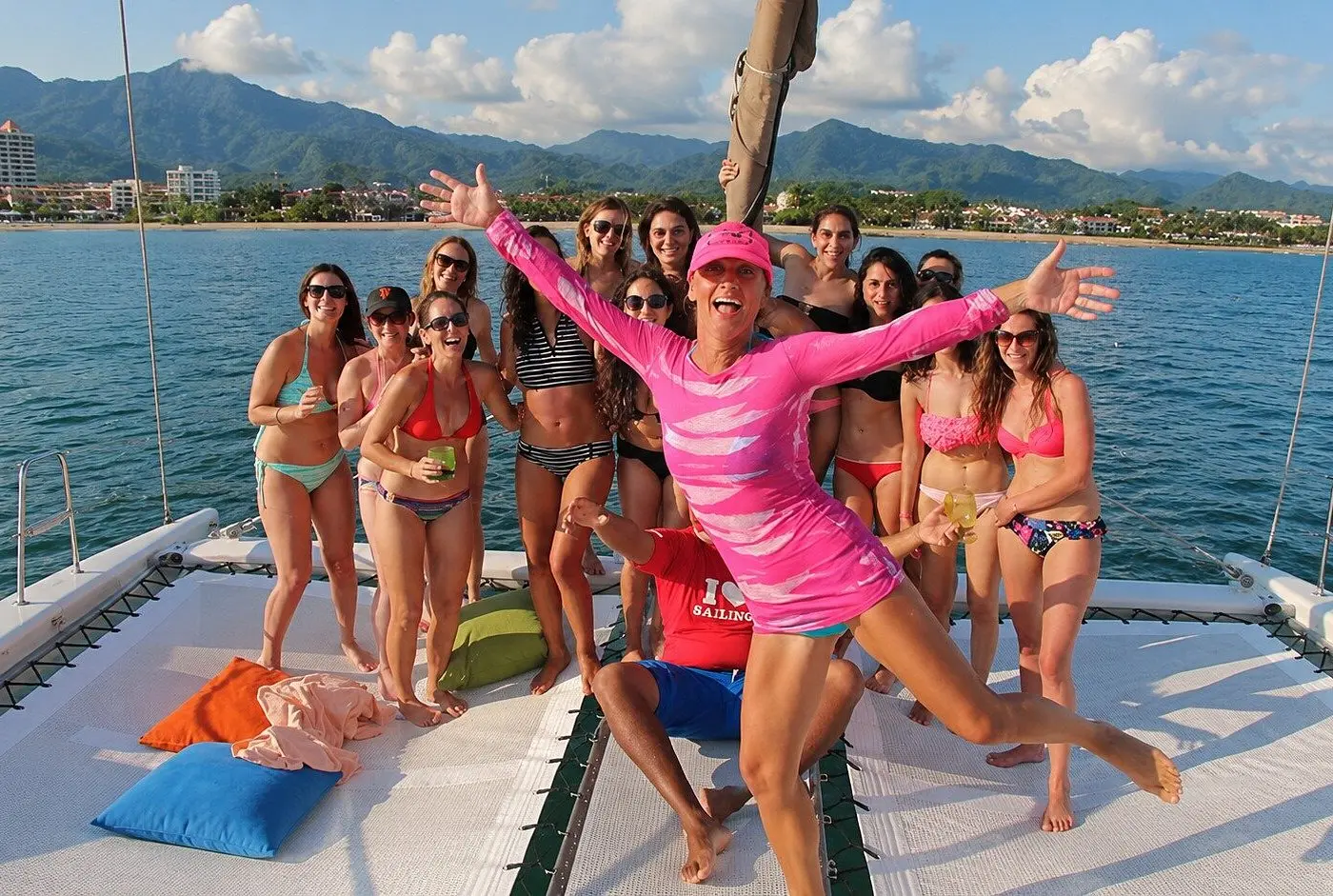 A group of women in swimsuits pose on the bow of a sailing catamaran with Puerto Vallarta's mountains behind