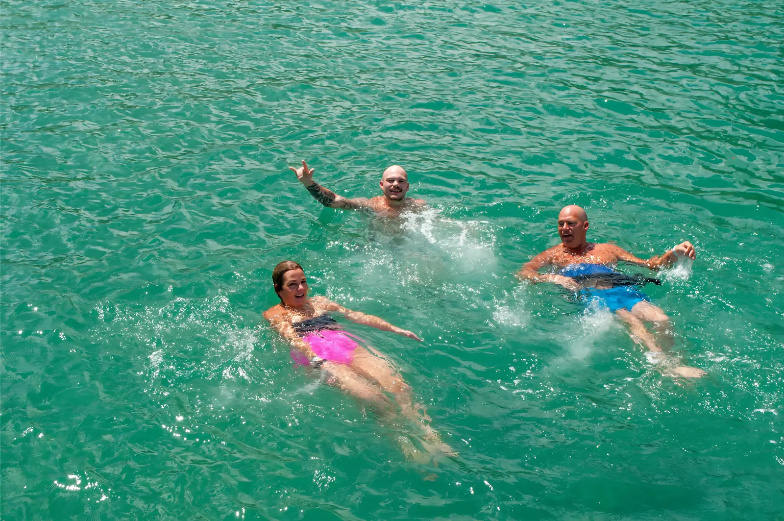 Three guests swimming and waving in clear turquoise water during a swim stop