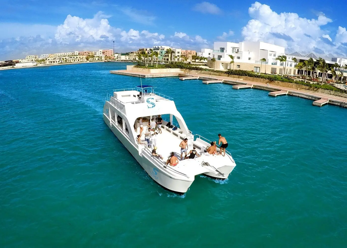 A double-deck catamaran with guests cruises through the Cap Cana marina past white villas and docks
