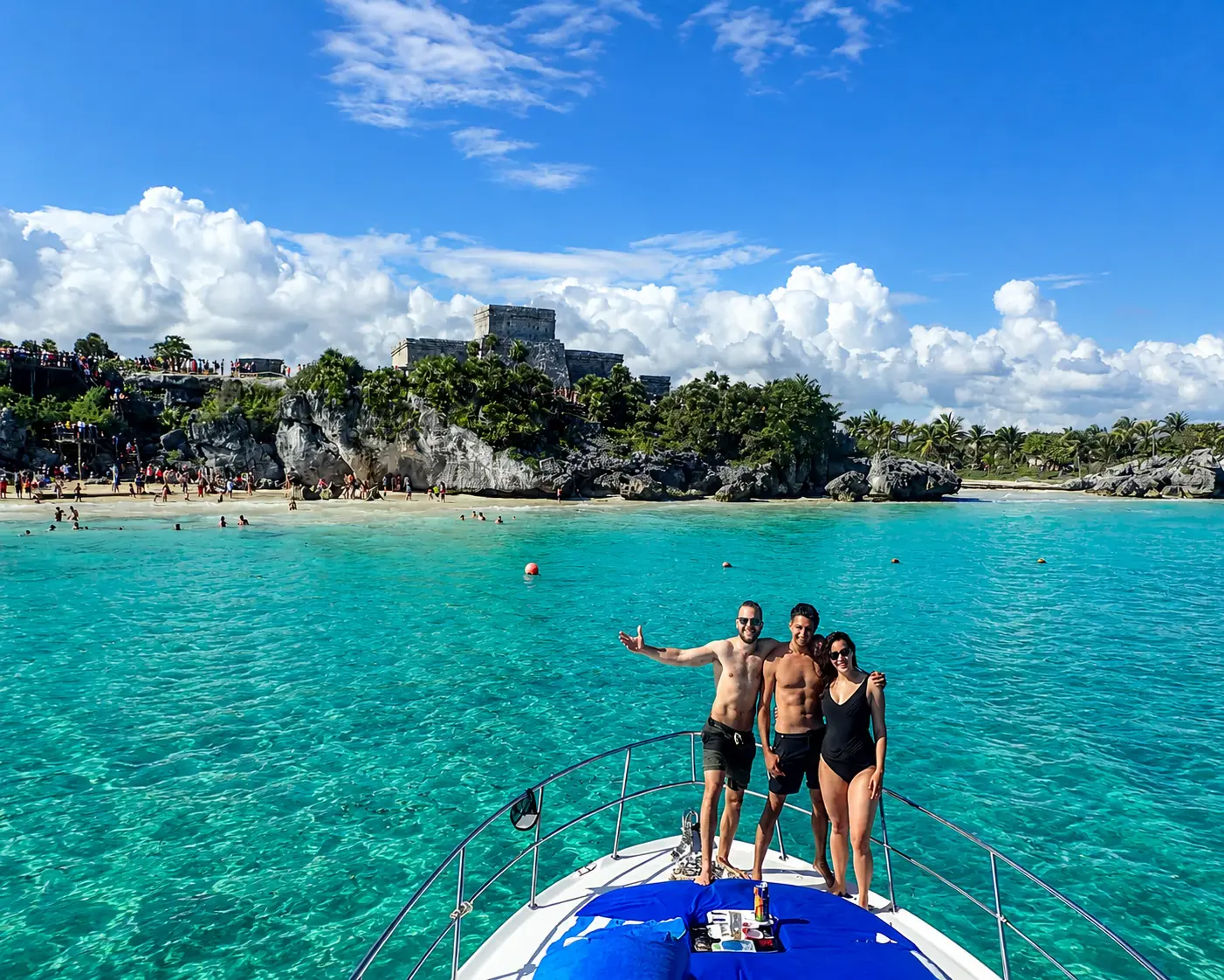 Three guests posing on the bow of a motor yacht in turquoise water below the clifftop Tulum ruins