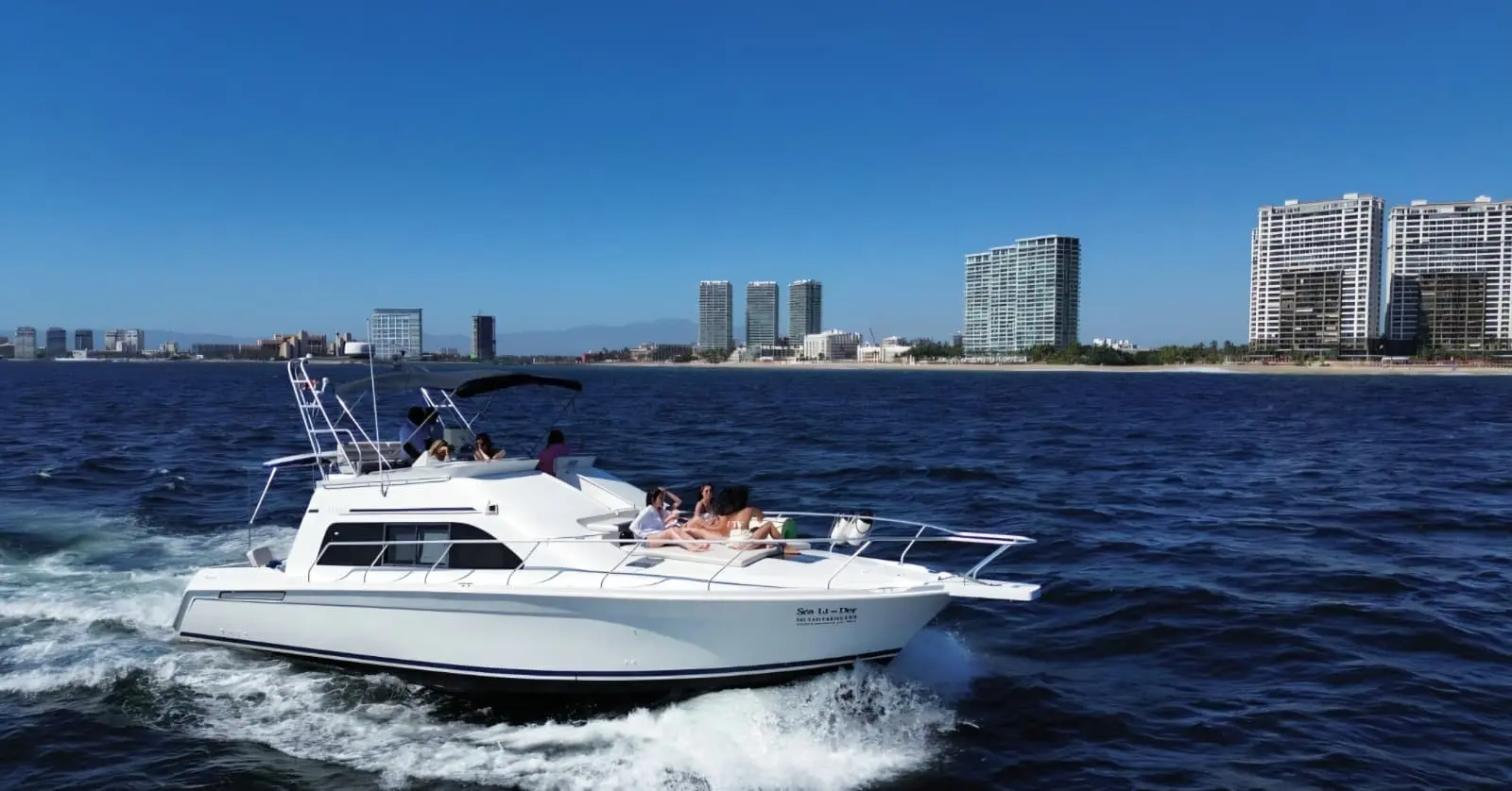 A flybridge yacht cruises past Puerto Vallarta's high-rise shoreline with guests sunning on the bow