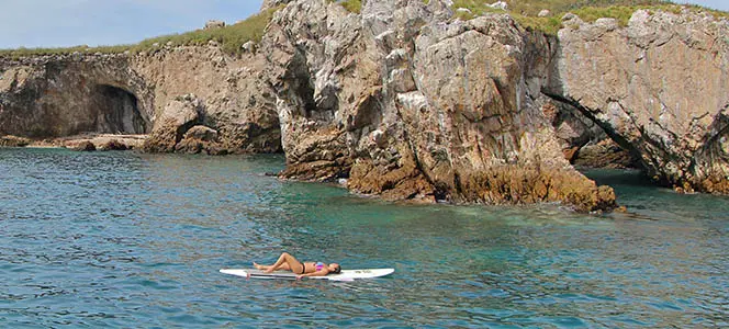 Woman sunbathing on a paddleboard beside rocky island cliffs and sea caves off Puerto Vallarta