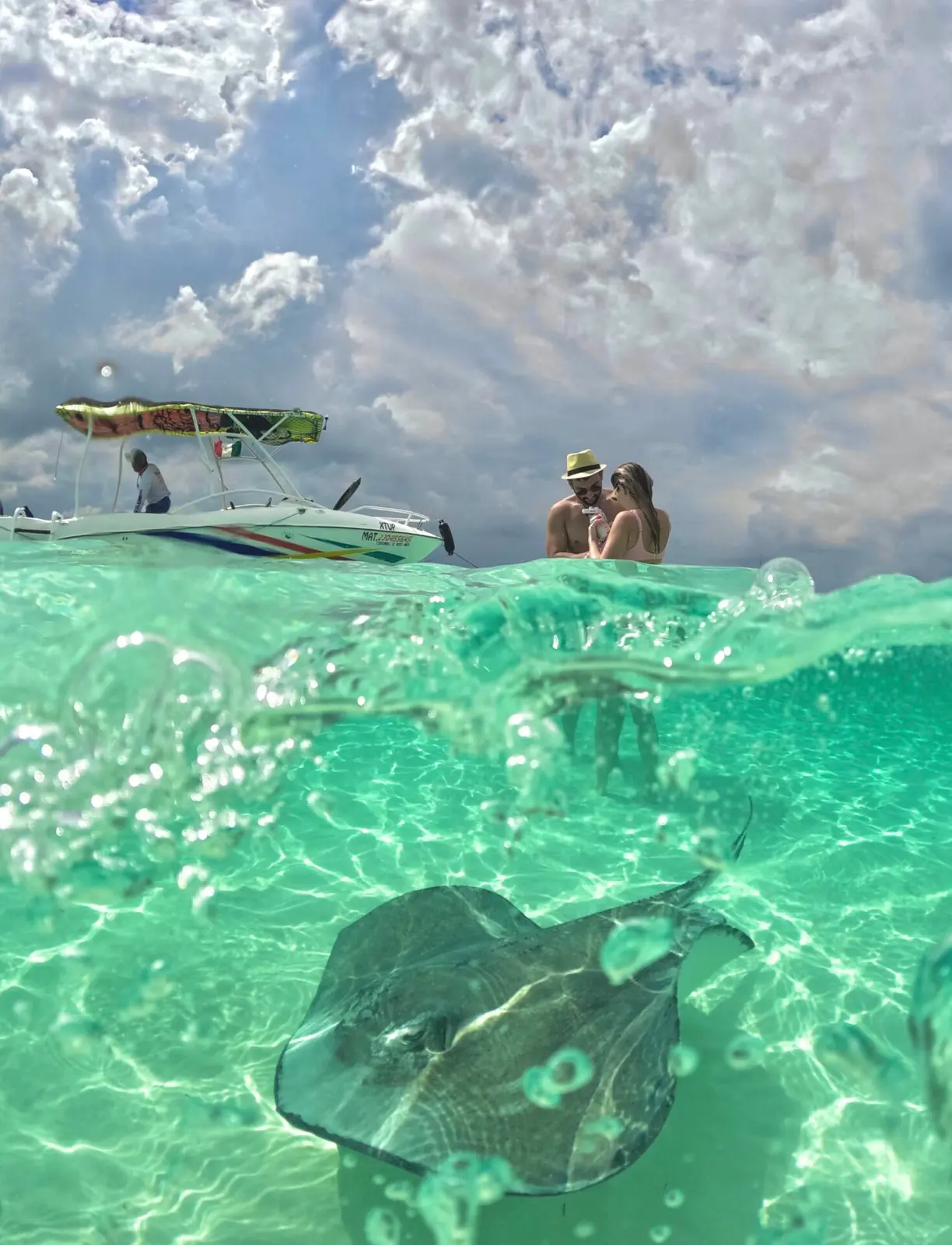 Over-under view of a stingray in clear shallow water with a couple and a speedboat above the surface