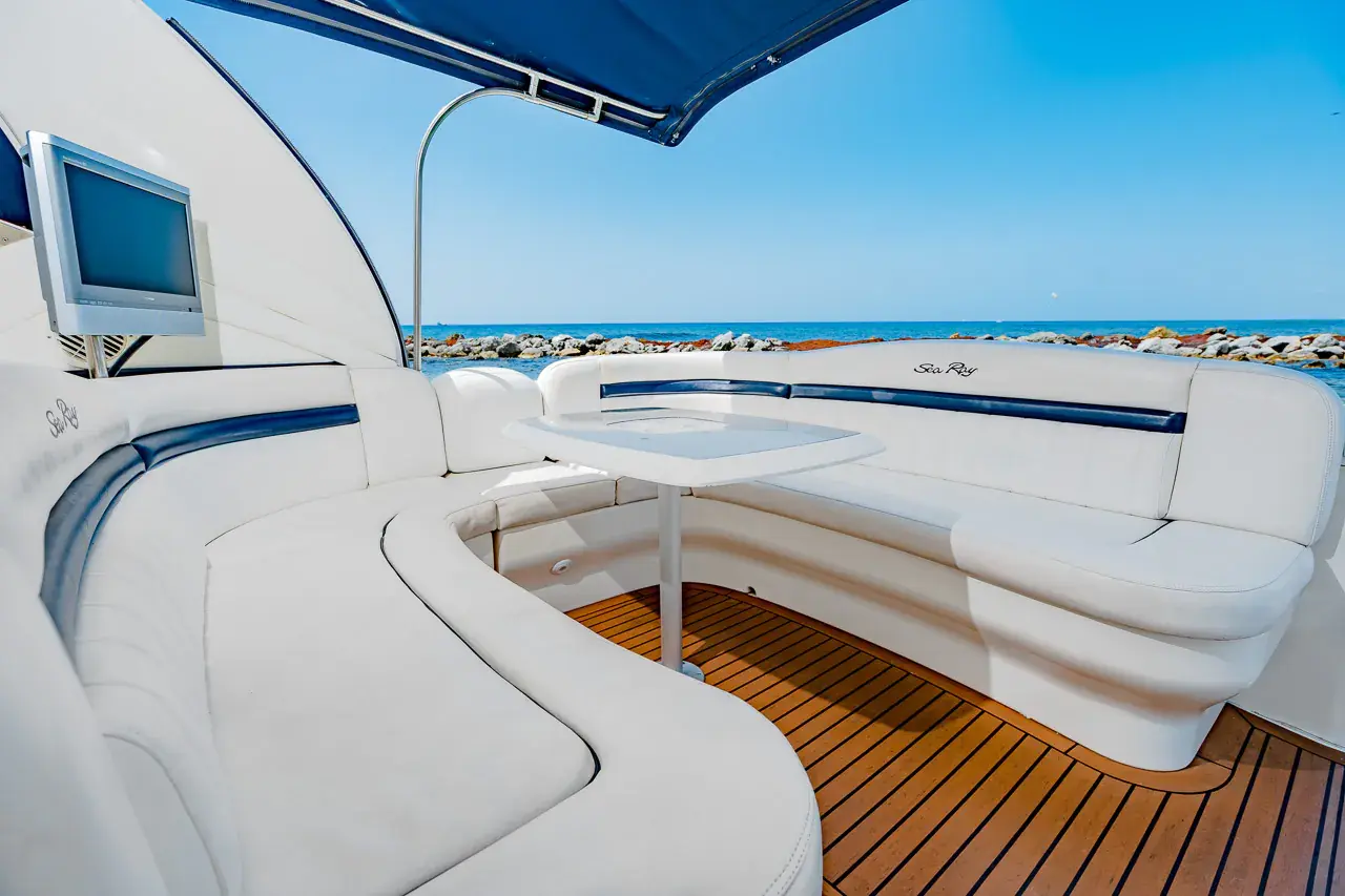 Shaded aft lounge of the yacht with curved white seating, a table, TV and teak floor under the bimini
