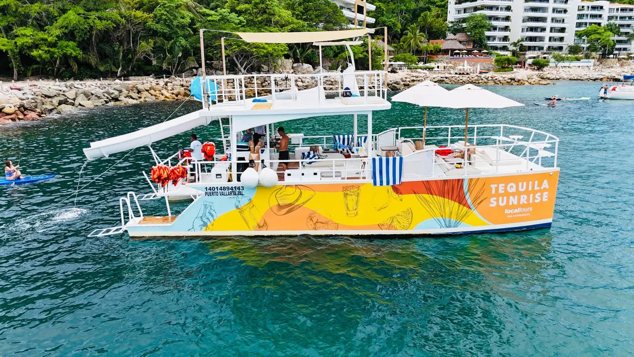 A colorful two-level party catamaran with a water slide anchored off a hotel-lined shore in Puerto Vallarta