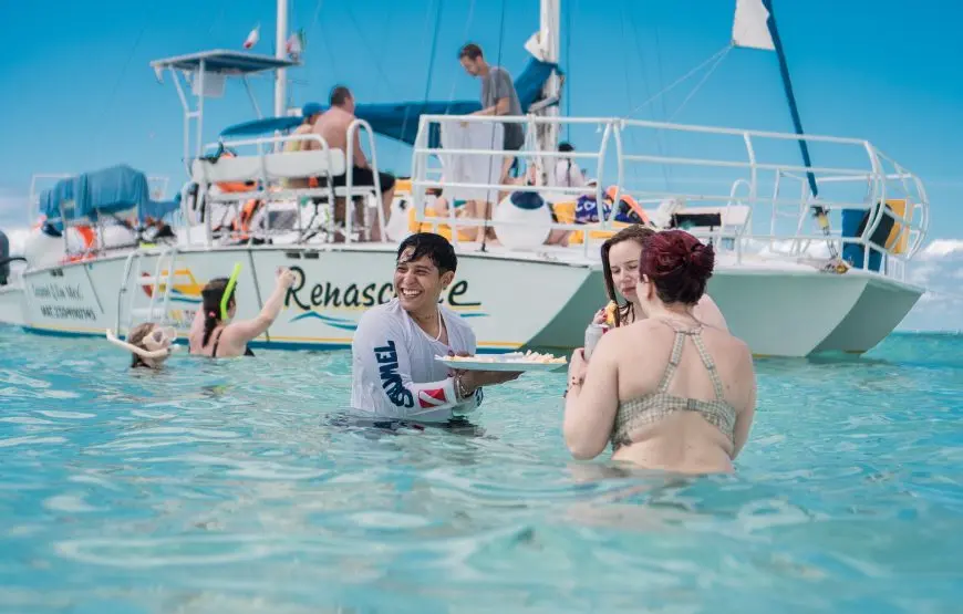 A crew member carries a tray to guests standing in the shallows beside the catamaran off Cozumel