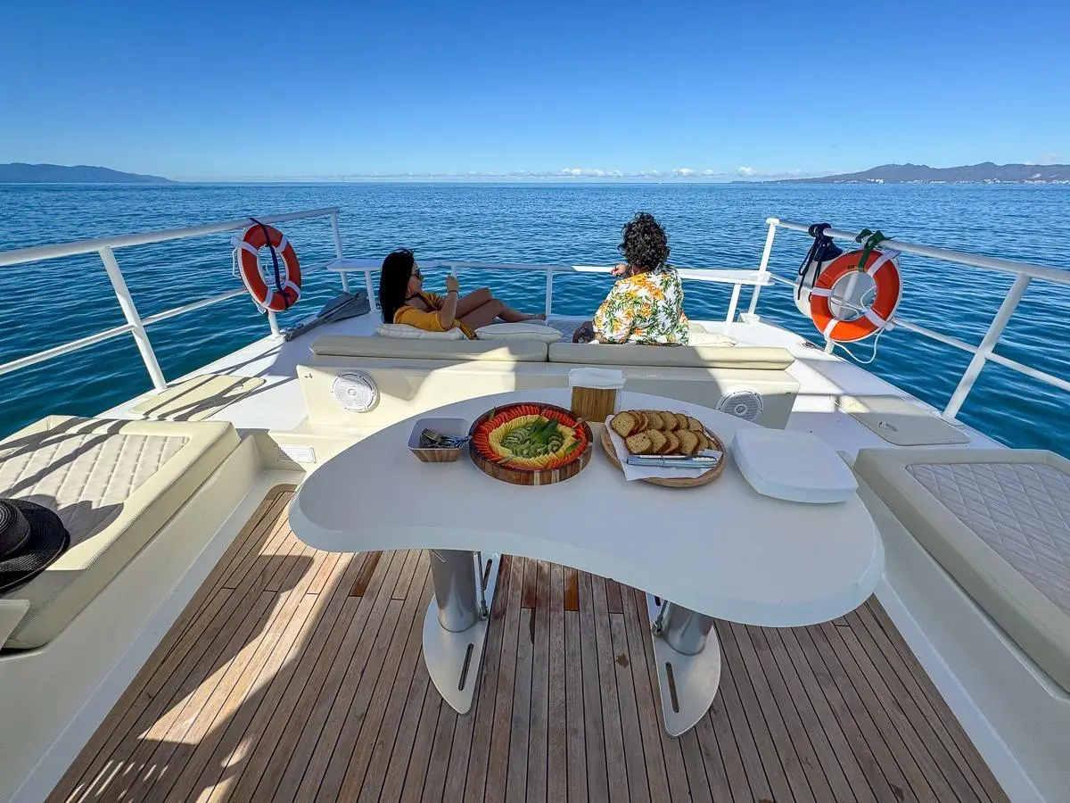 Two guests sit at the bow rail past a curved white table set with guacamole and chips on teak decking