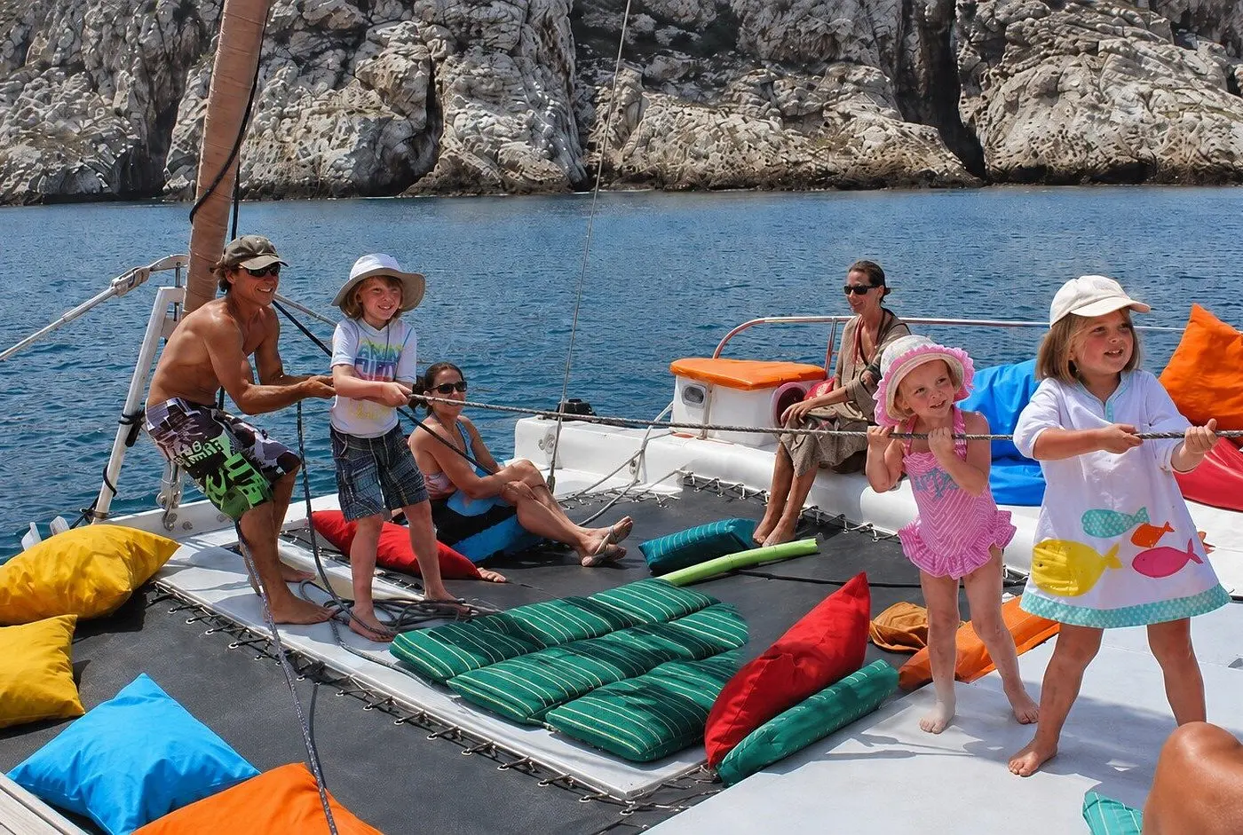 Kids and adults gather on the catamaran's netting among colorful cushions beside rocky cliffs