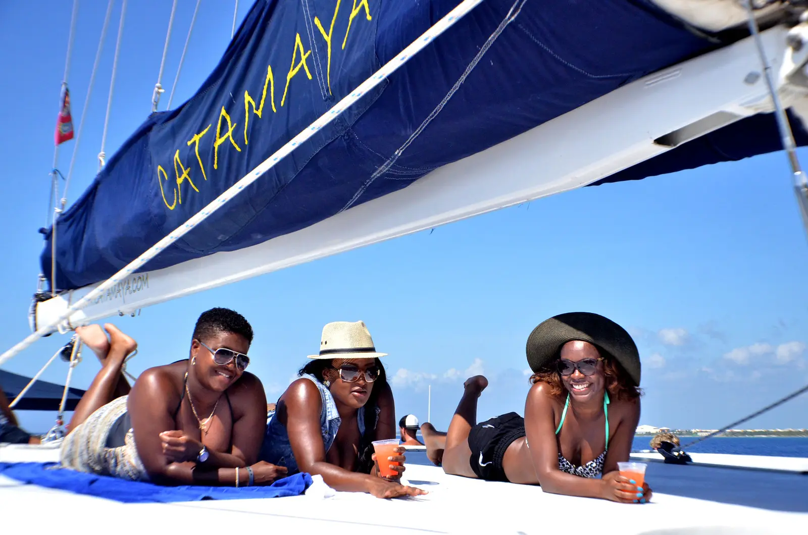 Three guests sunbathing on a blue towel on the catamaran's deck, drinks in hand under the sail cover
