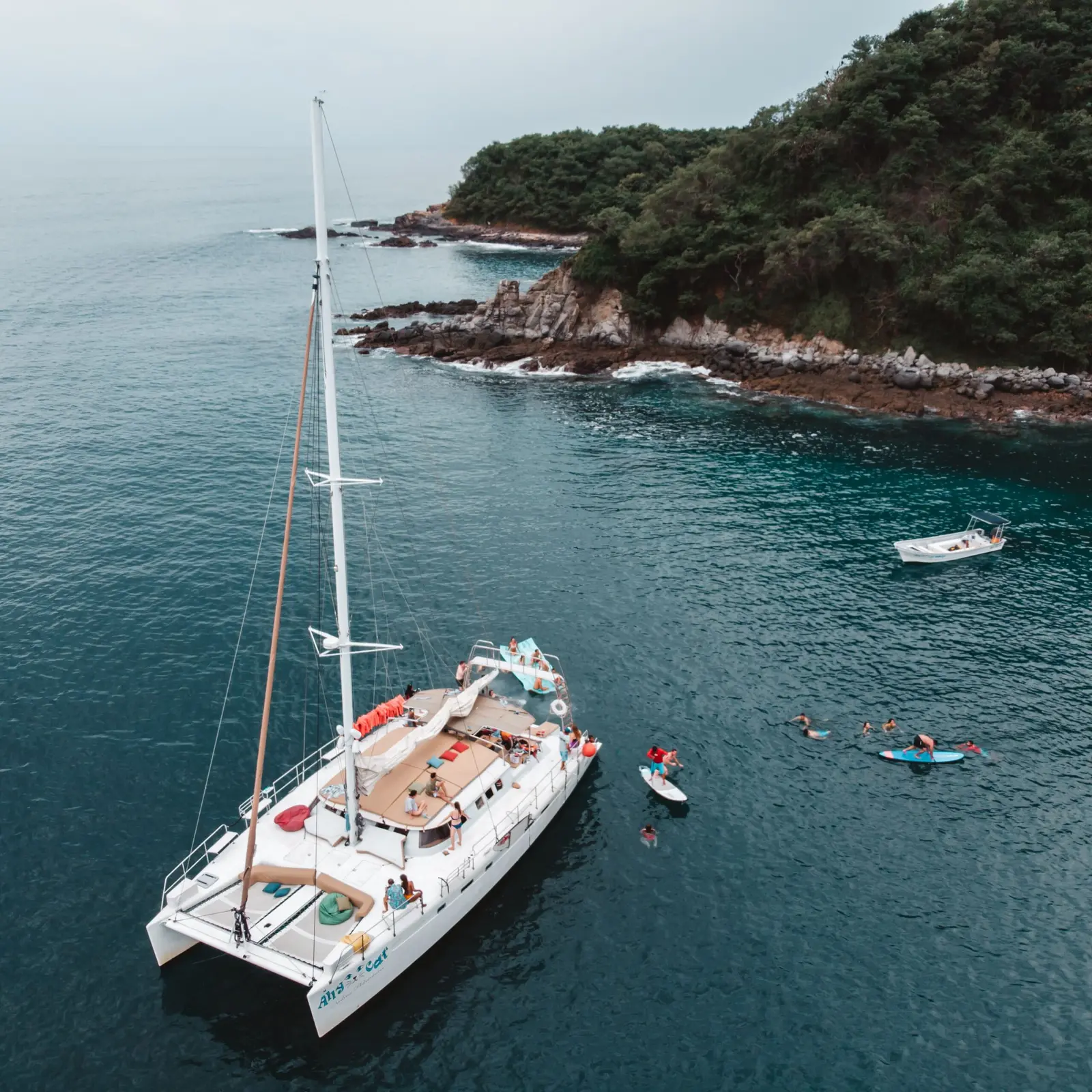 Aerial view of a sailing catamaran anchored off a forested headland with a dinghy and paddleboarders nearby