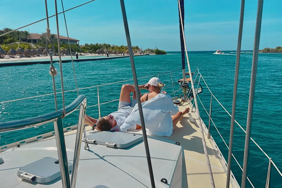 Couple stretched out on the foredeck of a sailing yacht passing a palm-fringed shore in Curaçao