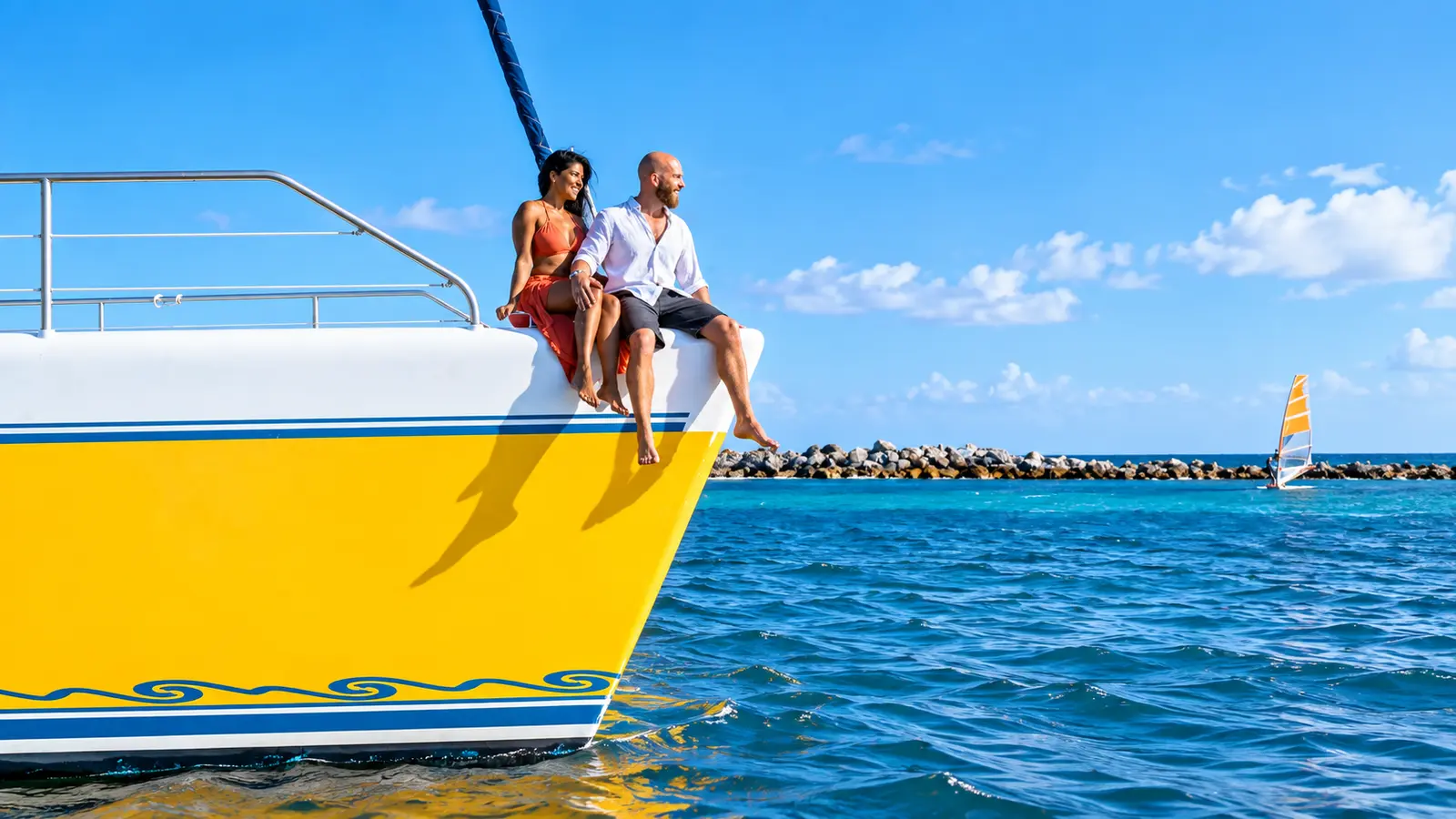 Couple sitting on the yellow bow of the catamaran above blue water near a Riviera Maya breakwater