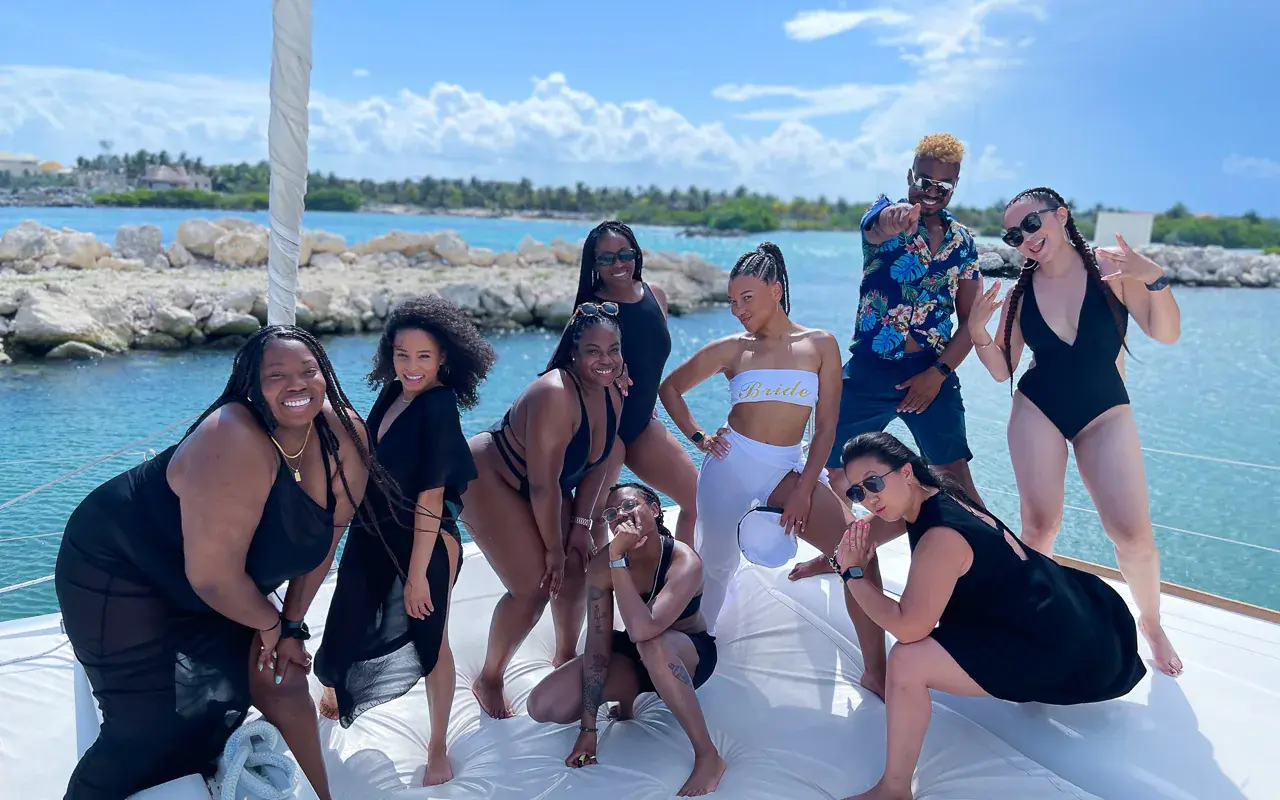 Friends in swimwear striking poses on the boat's bow pad by a rocky Riviera Maya breakwater