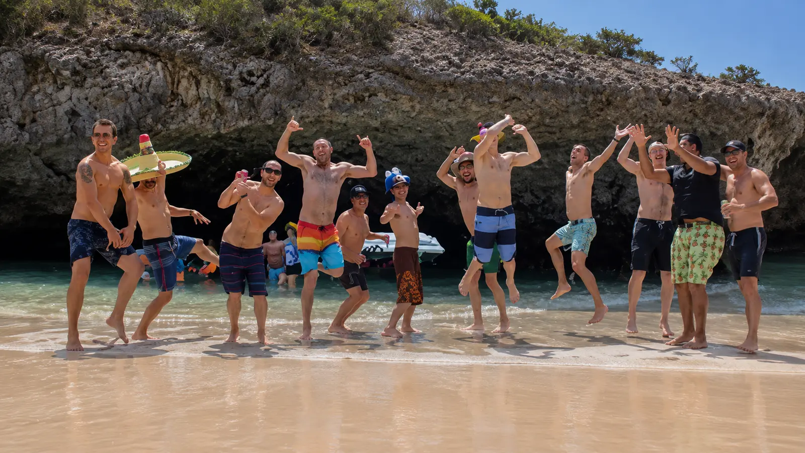 A group of men strike playful poses on a sandy beach in front of a rocky cave wall near Puerto Vallarta